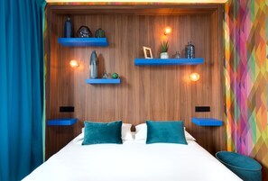 Una habitación de hotel moderna con cabecera de madera, almohadas color turquesa y estantes azules con objetos decorativos.