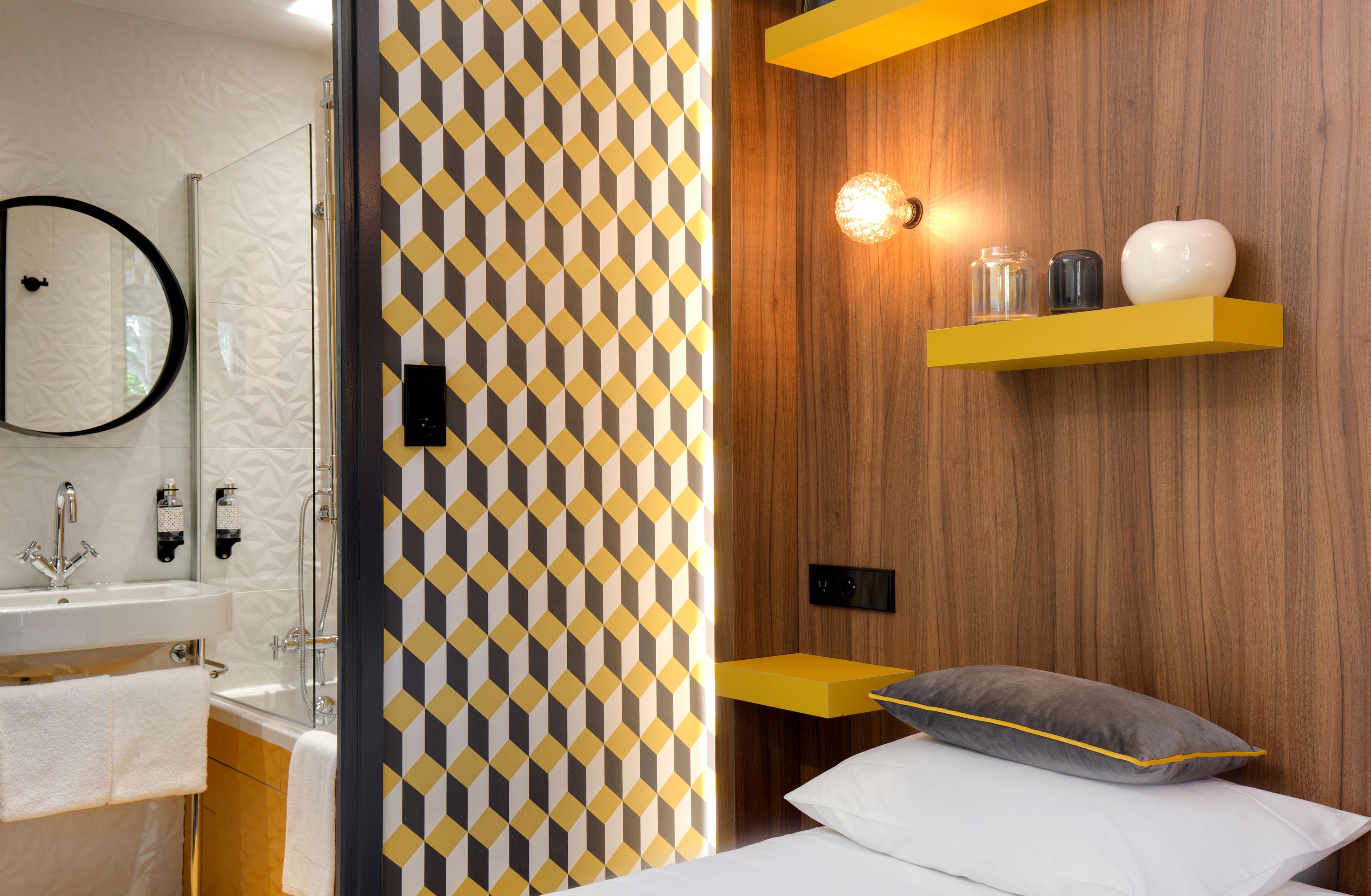 Un baño moderno con un revestimiento de pared de diseño geométrico, un panel de pared de madera y una mesita de noche con una lámpara y objetos decorativos.