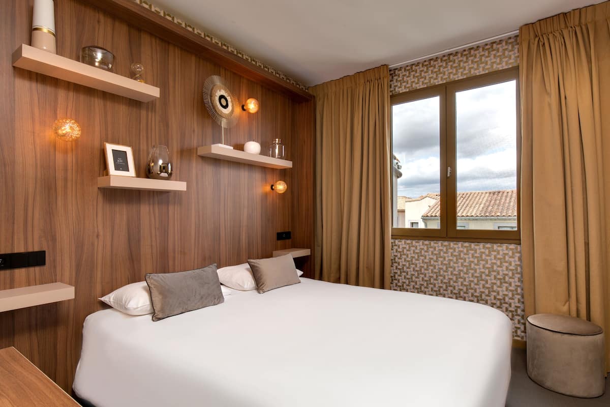 Ein Schlafzimmer mit einem großen Bett, Holzvertäfelung, eingebauten Regalen mit Dekoration und einem Fenster mit Vorhängen, mit Blick auf Gebäude.