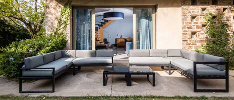 Un espace extérieur moderne aménagé avec un canapé d’angle et une table basse, un escalier menant à un niveau supérieur et un mur en pierre.