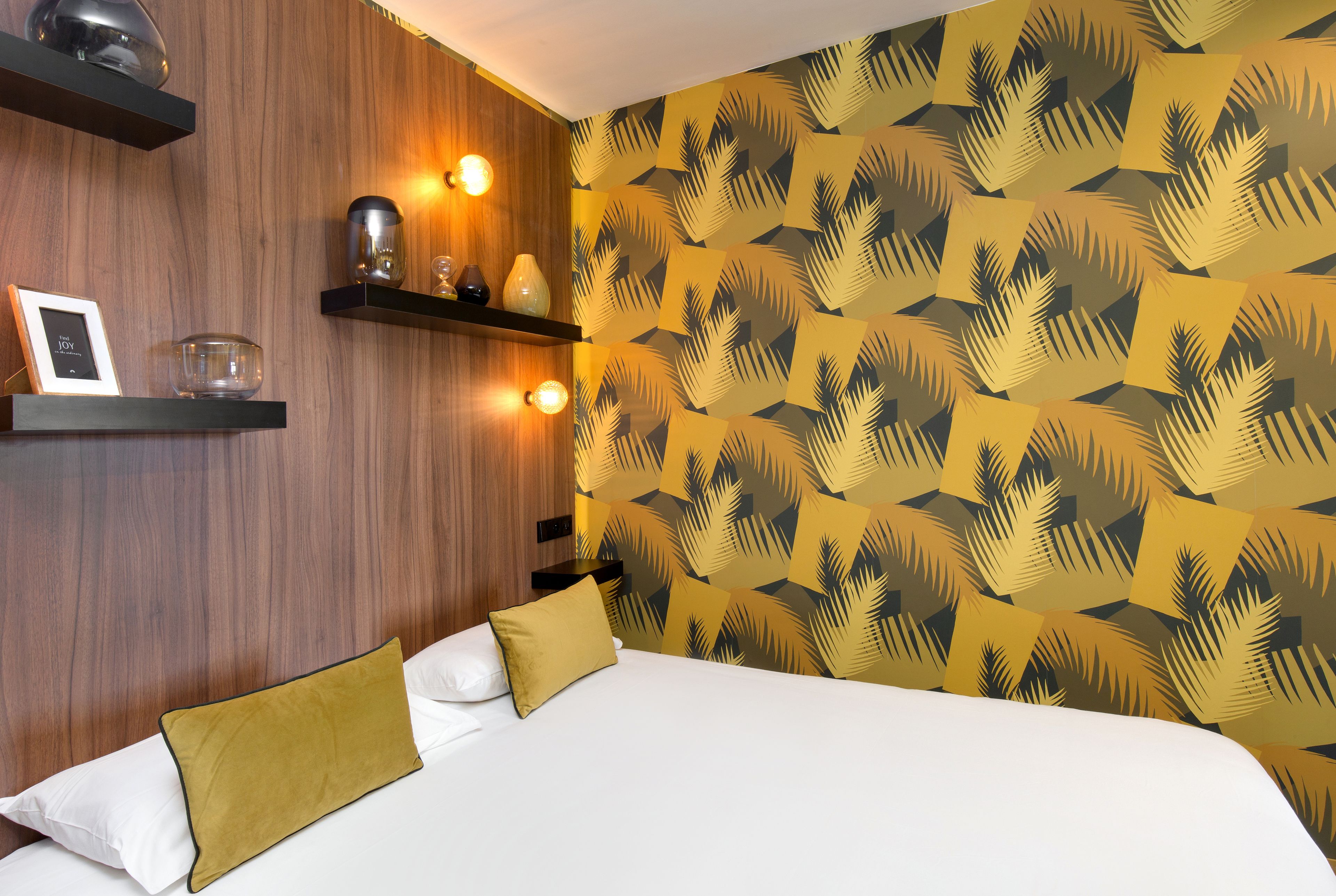 Un dormitorio con cabecera de madera, pared estampada y mesitas de noche con velas y un marco de fotos.