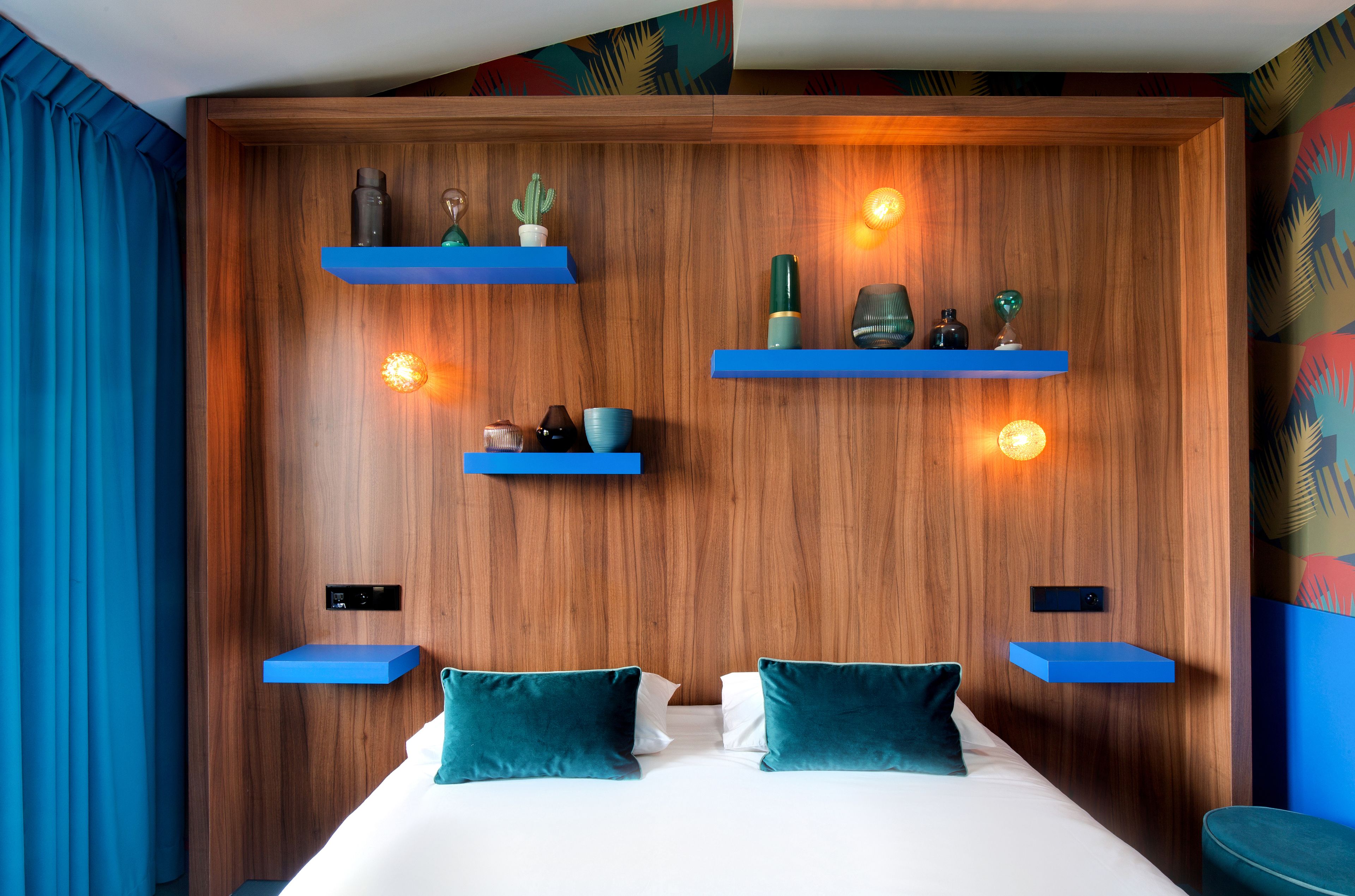 A modern hotel room with a wooden headboard, blue shelves with decorative items, and a bed with teal pillows.