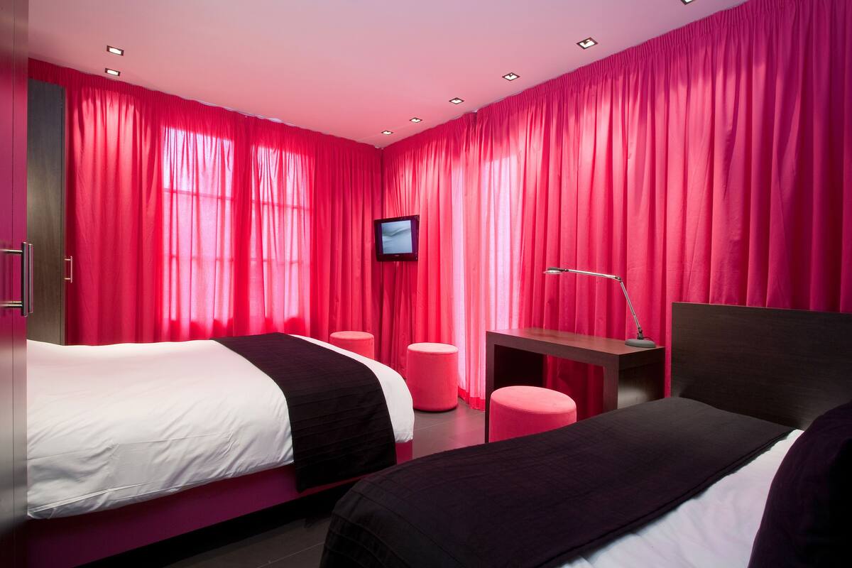Ein modernes Hotelzimmer mit zwei Betten, einem Flachbildfernseher, einem Schreibtisch mit Lampe und pinkfarbenen Vorhängen.