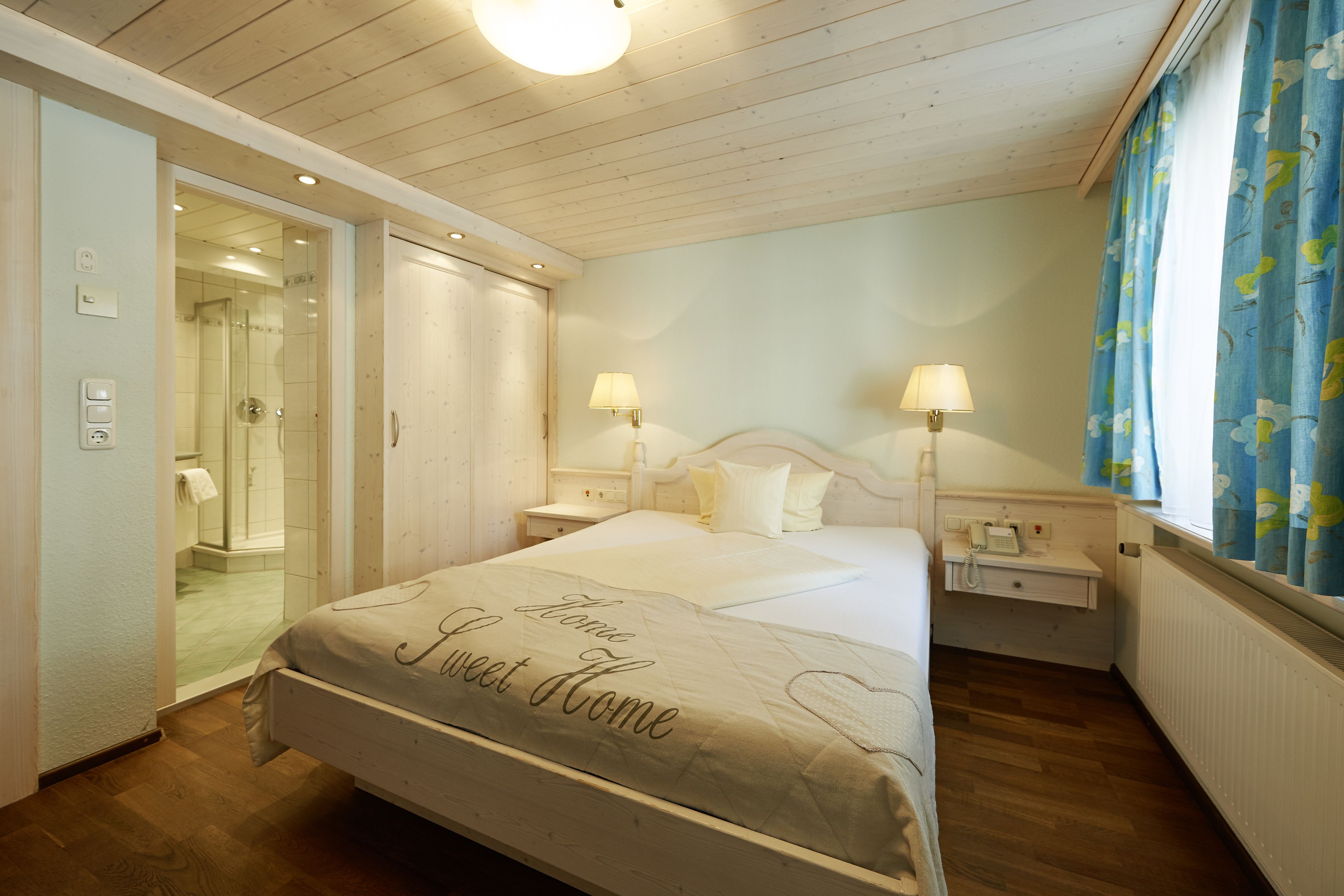 Un dormitorio con cama, mesitas de noche, lámpara, ventana con cortinas, puerta que da a un baño y techo de madera.