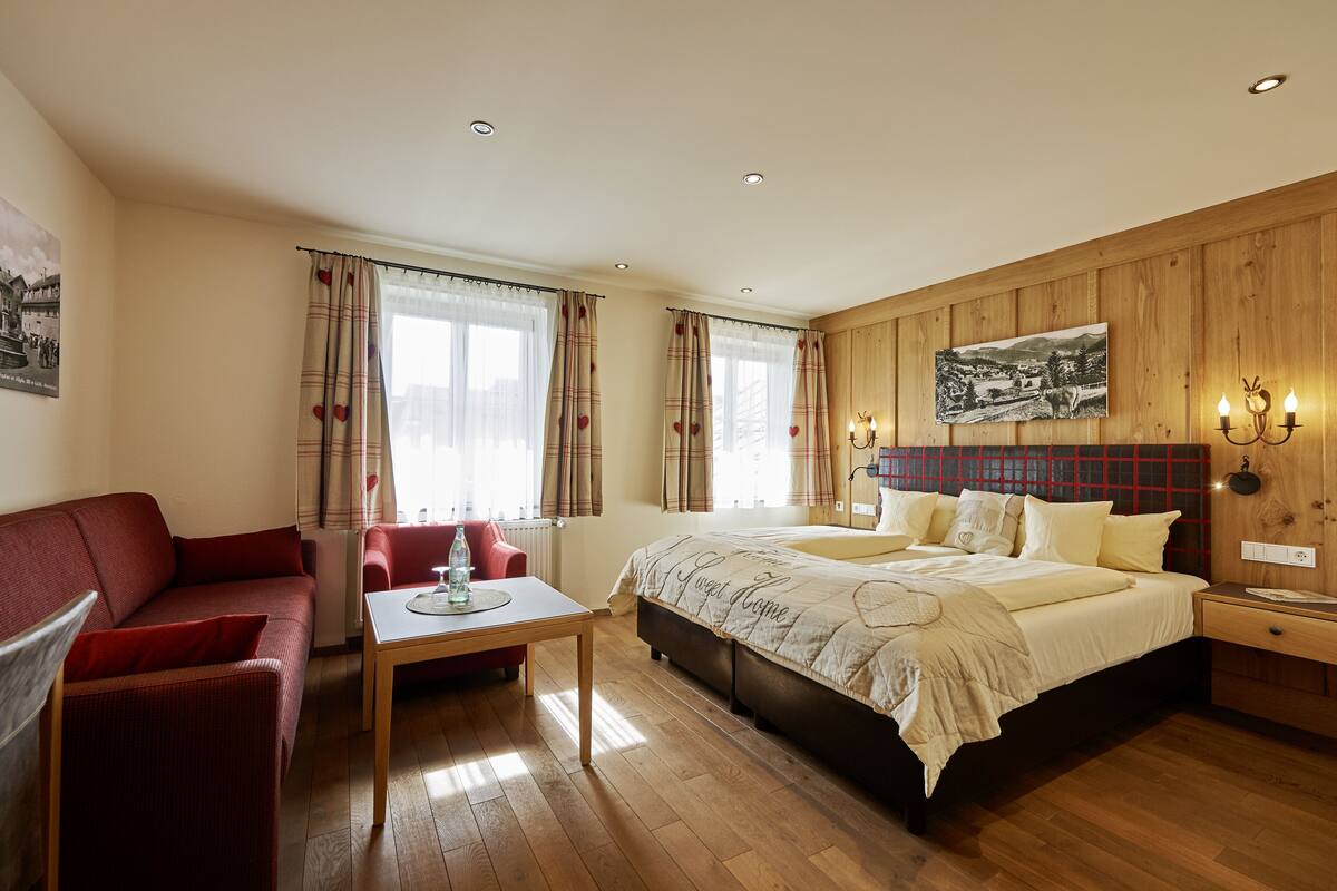A bedroom with a bed, a red sofa, a wooden table, and a window with curtains.
