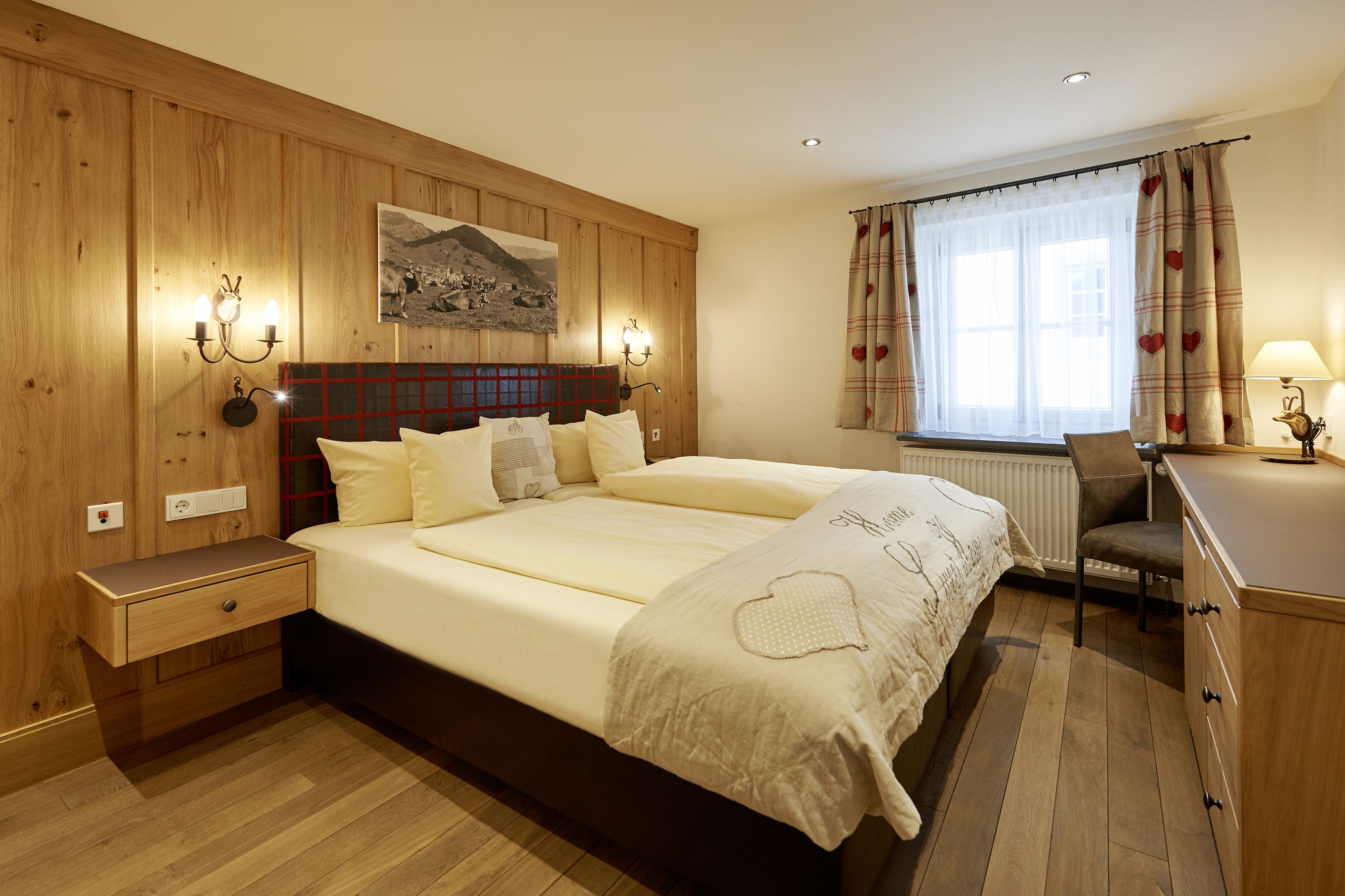 A bedroom with a large bed, wooden panel walls, a desk with a chair, and a window with curtains.