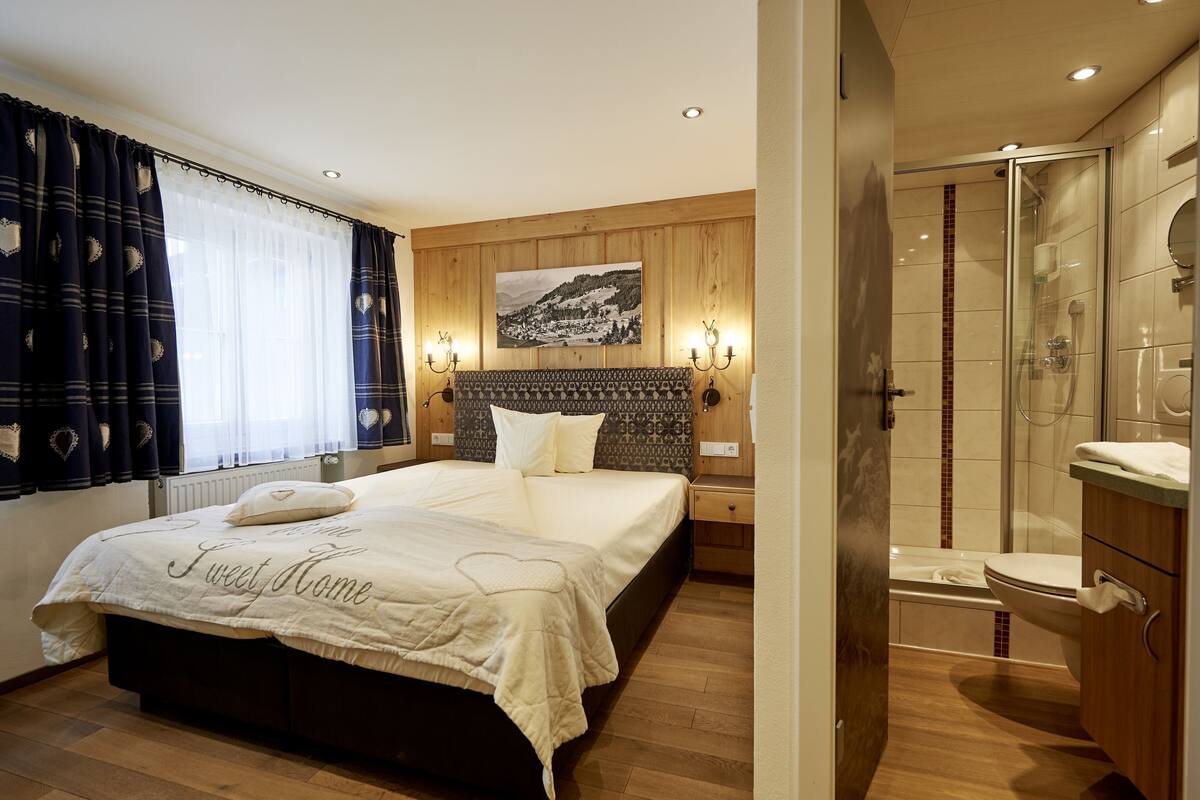 A bedroom with a bed, a nightstand, a headboard, a window with curtains, a wall-mounted picture, and a bathroom with a shower, toilet, and sink.