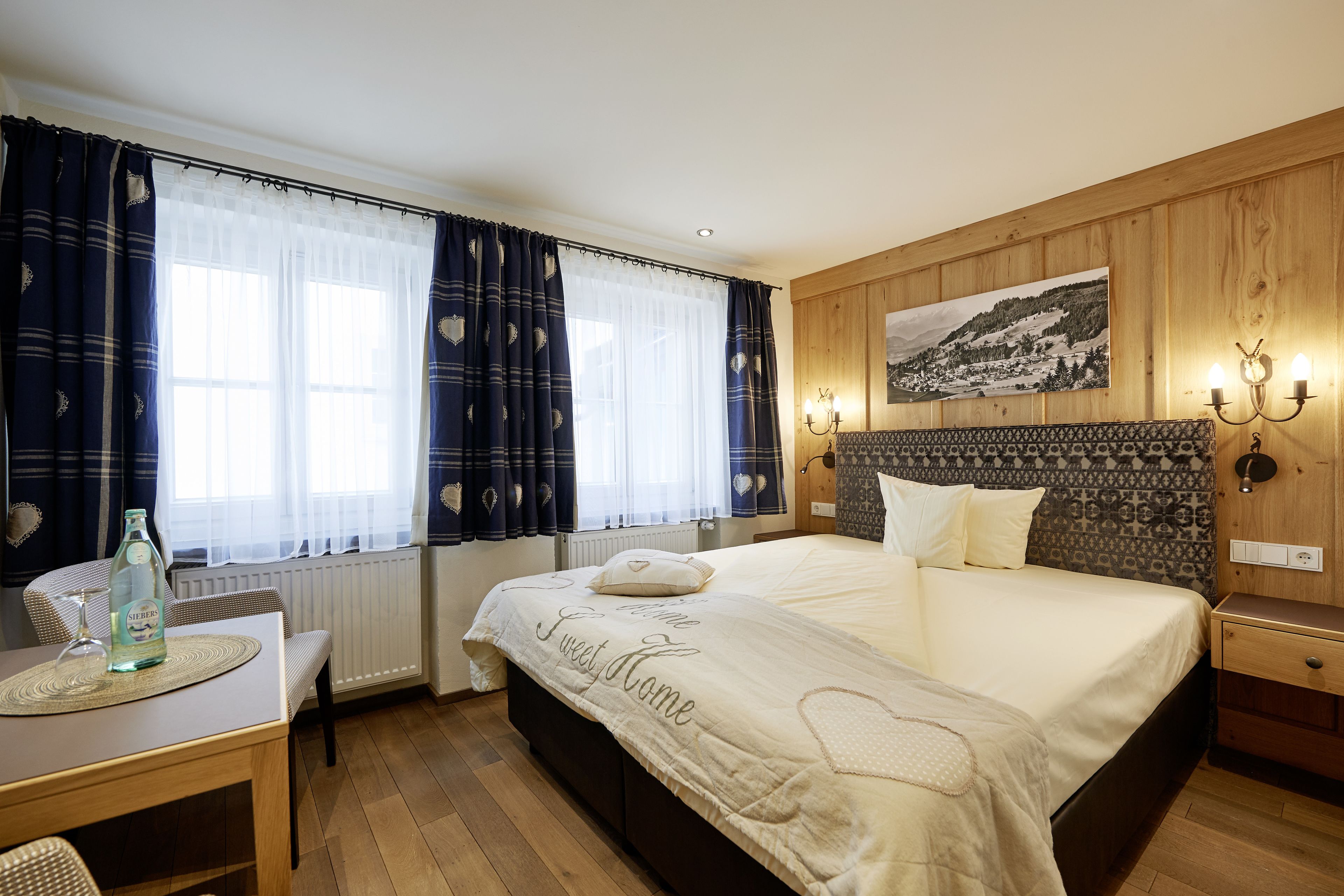 A bedroom with a large bed, a wooden headboard, a bedside table, a chair, a table with a bottle and glass, and a window with curtains.