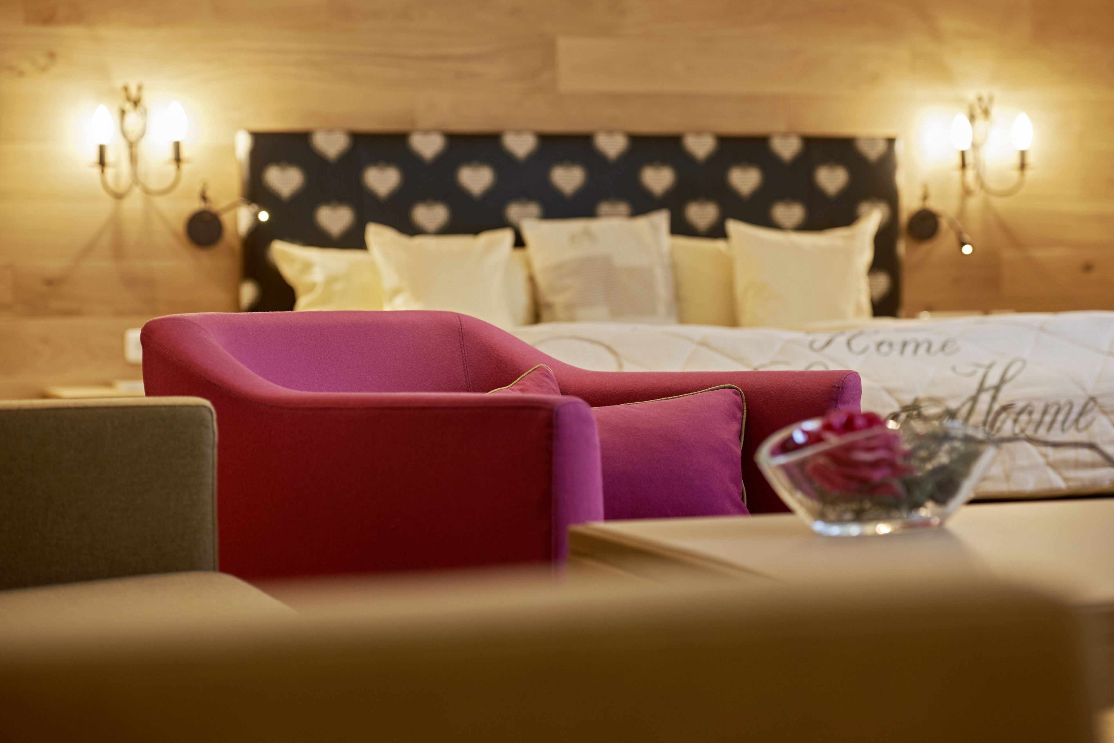 Un dormitorio con cama, una silla roja, una mesita de noche con un florero y un papel tapiz con diseño de corazones.