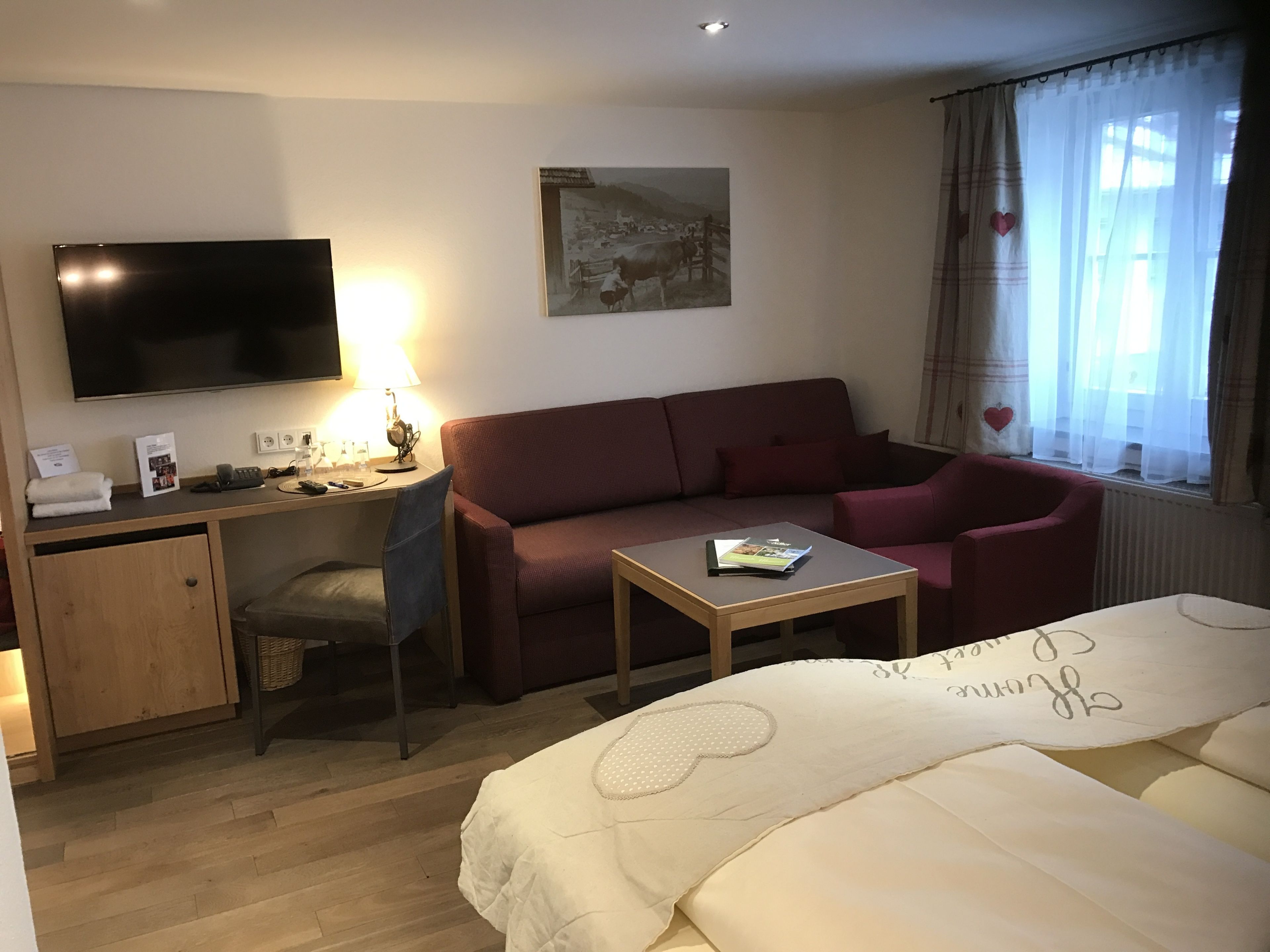 Ein Hotelzimmer mit Bett, Schreibtisch mit Computer, Sofa, Fernseher und Fenster mit Vorhängen.