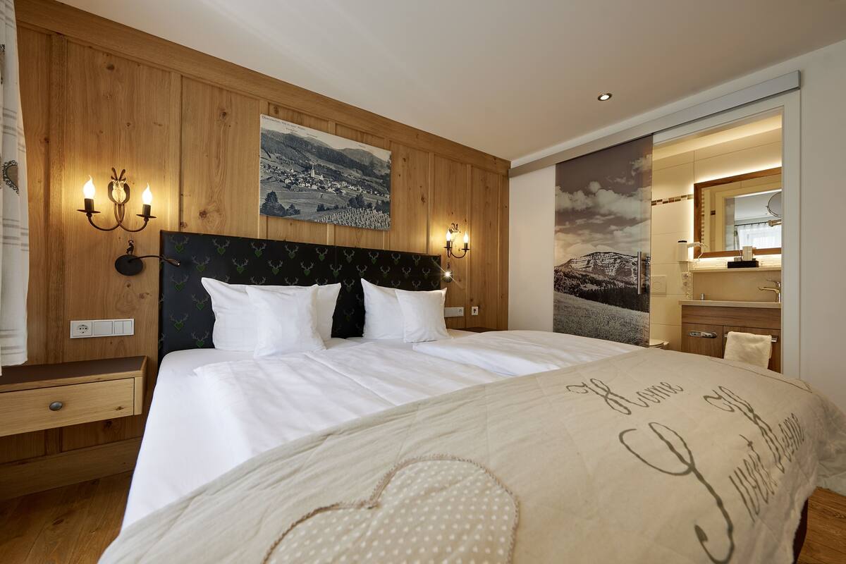 A hotel room with a wooden headboard, a bed with white linens, a nightstand, a wall-mounted lamp, and a painting of a landscape.