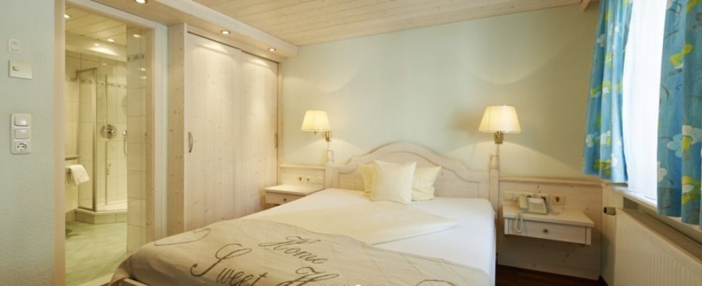A hotel room with a bed, two bedside lamps, a nightstand, a window with curtains, and a door leading to a bathroom.