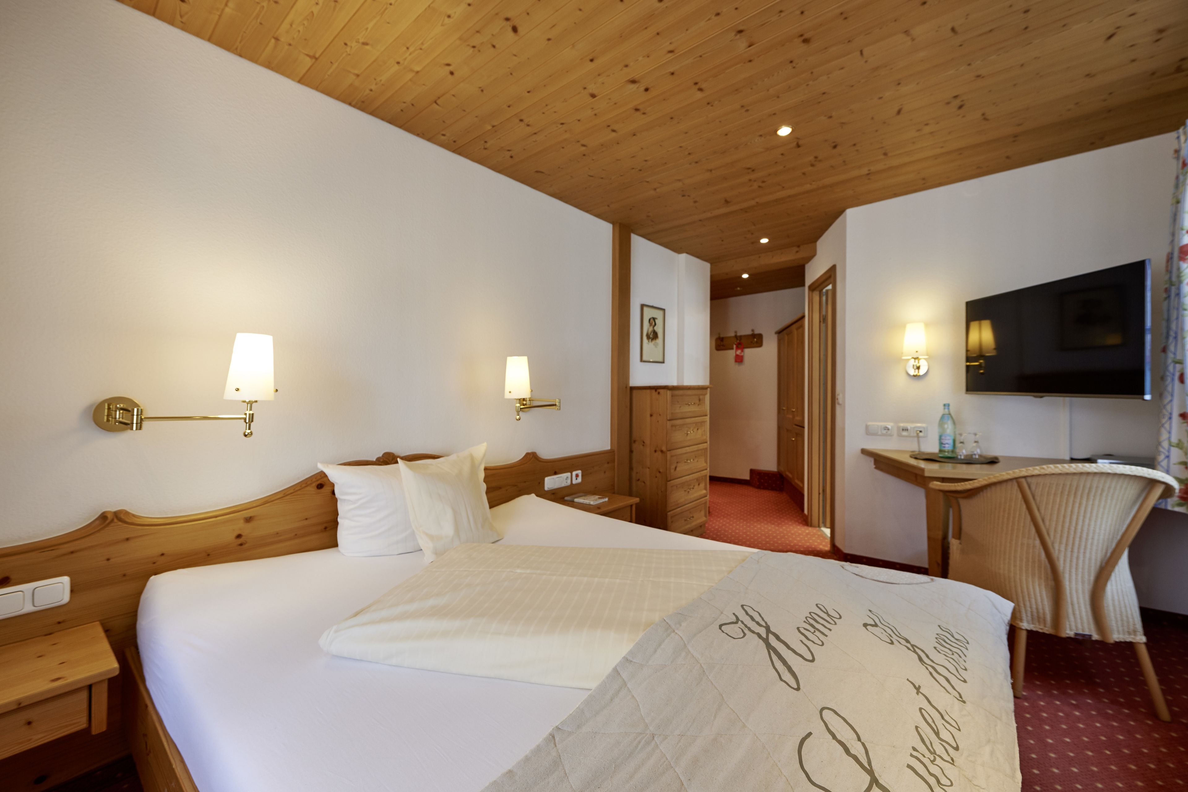 A hotel room with a wooden ceiling, a bed with white linens, a bedside table with a lamp, a desk with a chair, and a television on the wall.