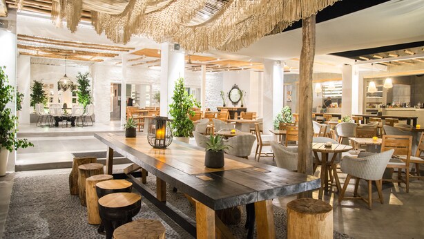 A spacious, well-lit indoor area with wooden tables and chairs, potted plants, and a rustic ceiling design. - Makris Beach Hotel (Santorini)