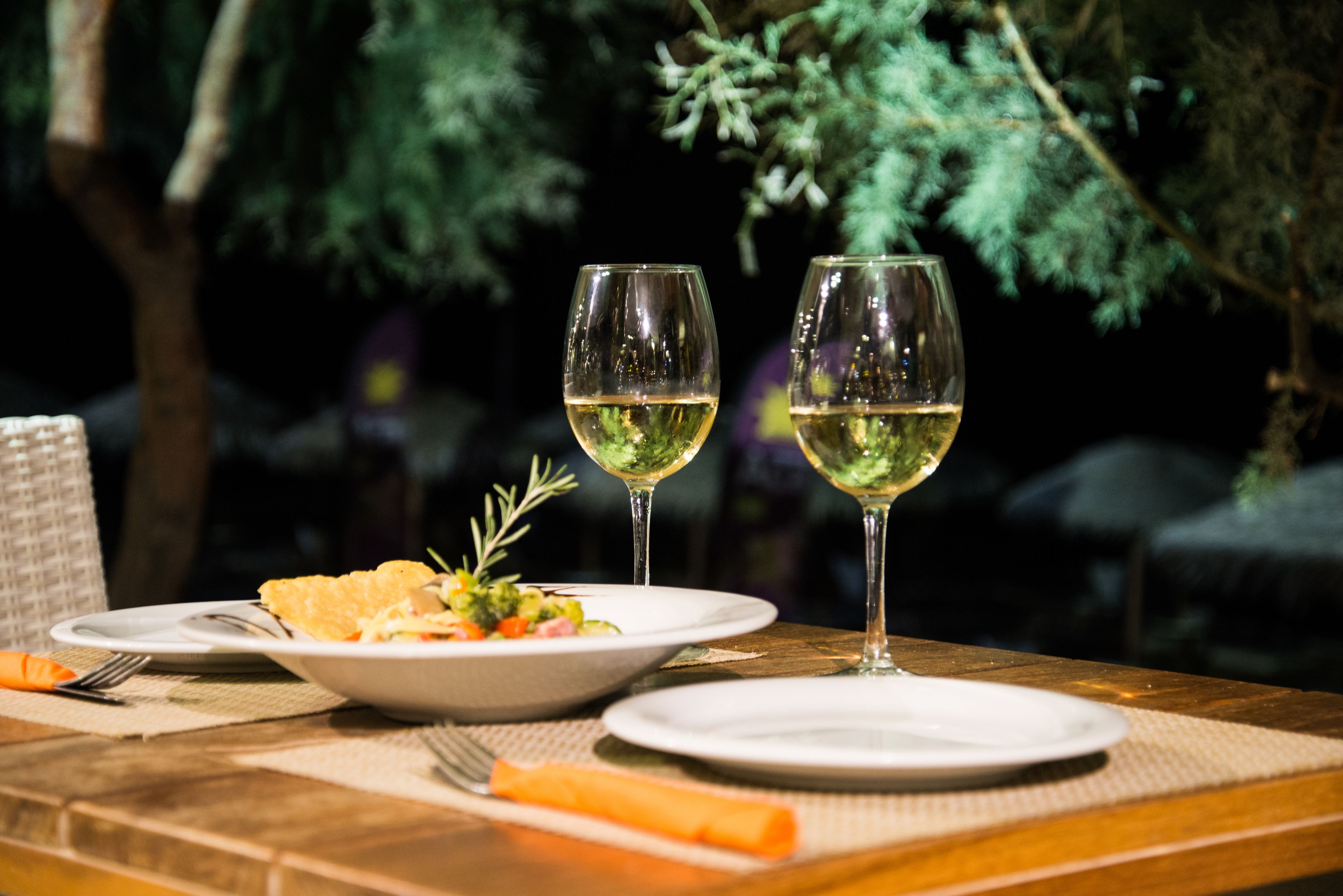 A table with plates of food and two glasses of white wine, set outdoors with trees in the background.
