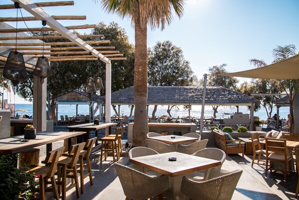An outdoor patio with wooden tables and wicker chairs, overlooking a beach. - Makris Beach Hotel (Santorini)