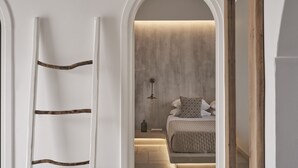 A bedroom with a bed, bedside table, lamp, and a view through an arched doorway. - Makris Beach Hotel (Santorini)