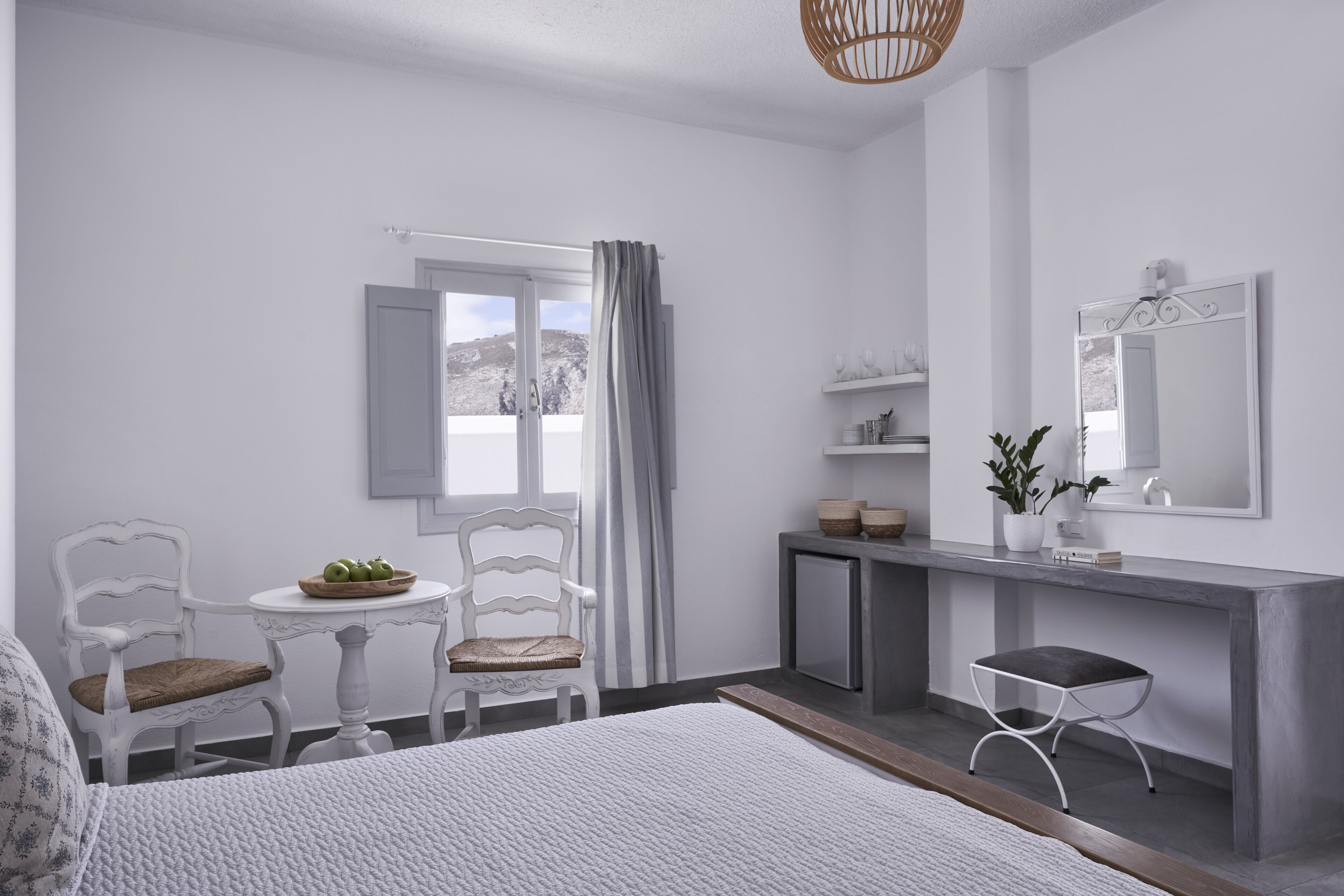 A hotel room with a bed, a desk, a chair, a small table with a bowl of fruit, a potted plant, and a window with curtains.