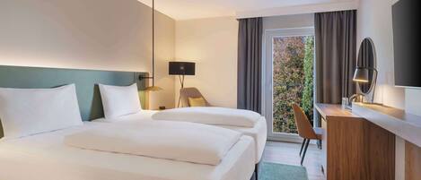 A hotel room with two beds, a desk, a chair, a TV, and a view of trees through the window.