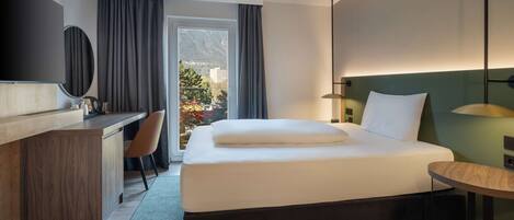Quarto de hotel com cama, escrivaninha, cadeira e vista para as montanhas pela janela.