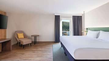 Habitación de hotel con cama, una silla, una mesita, televisor y vistas a un puente y un bosque.