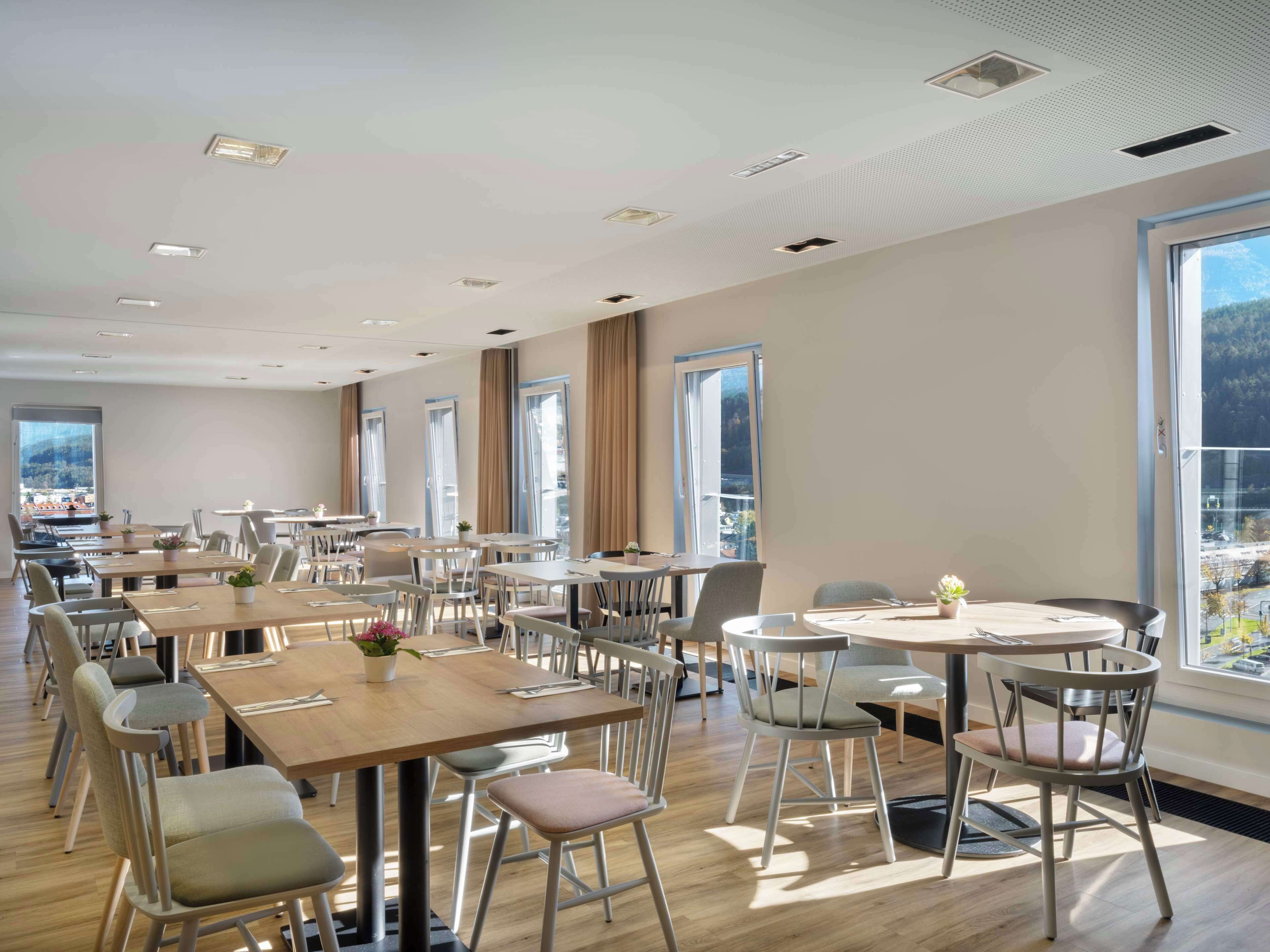 A spacious dining area with multiple tables and chairs, large windows offering a view of the outdoors, and a modern ceiling design.