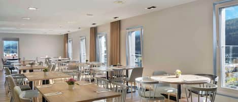 A spacious dining area with multiple tables and chairs, large windows offering a view of the outdoors, and a modern ceiling design.