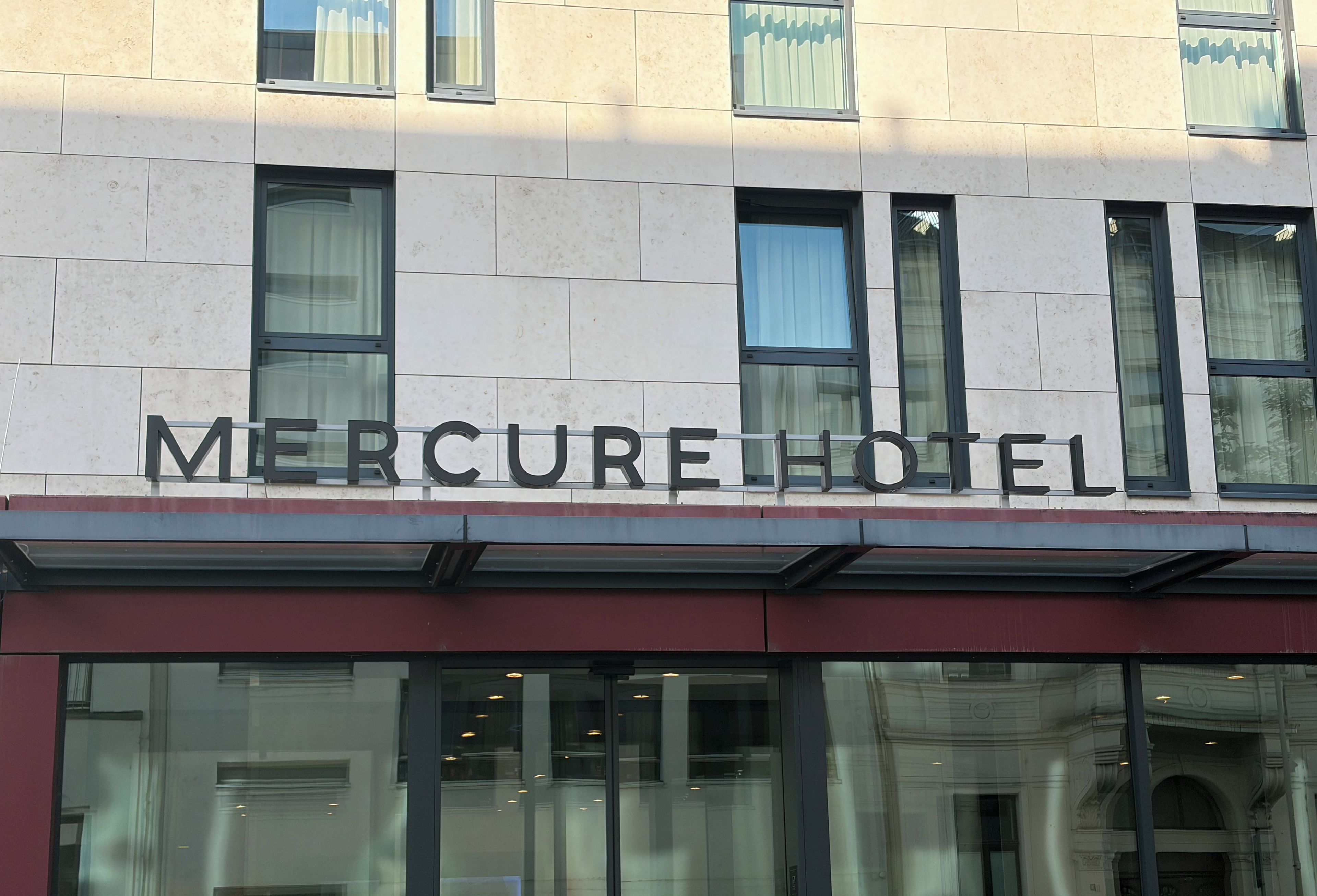 Une enseigne d’hôtel portant le nom « Mercure Hotel », au-dessus de la façade d’un bâtiment moderne doté de grandes fenêtres et d’un auvent rouge.