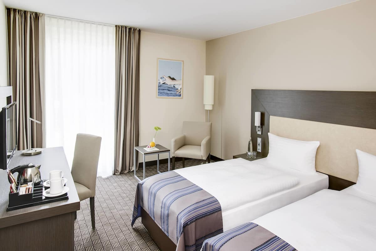 Ein Hotelzimmer mit zwei Betten, einem Schreibtisch, einem Stuhl, einem kleinen Tisch mit einer Vase und einem Bild an der Wand.