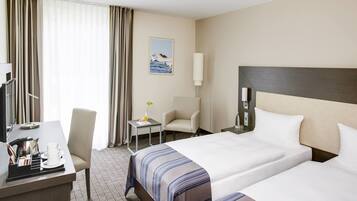 A hotel room with two beds, a desk, a chair, a small table with a vase, and a painting on the wall.
