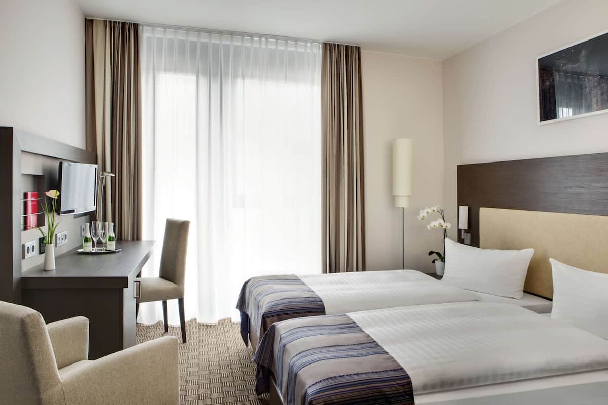 Ein Hotelzimmer mit zwei Betten, einem Schreibtisch mit Stuhl, einem Fernseher und einem großen Fenster mit Vorhängen.