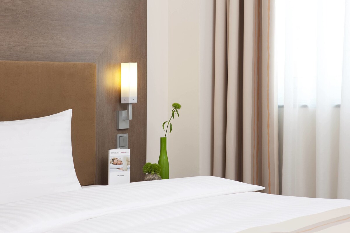A hotel room with a large bed, a nightstand with a lamp, a small potted plant, and a view of the outdoors through sheer curtains.