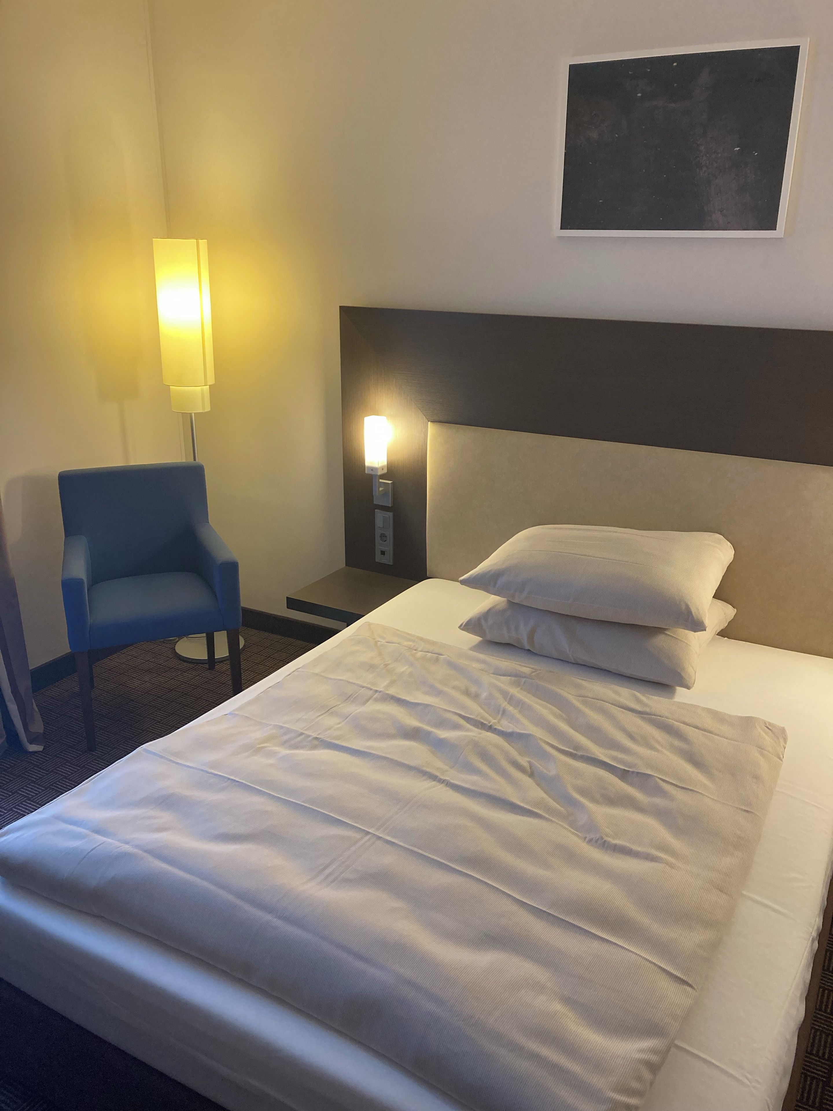 A hotel room with a bed, a chair, a nightstand, and a wall-mounted light fixture.