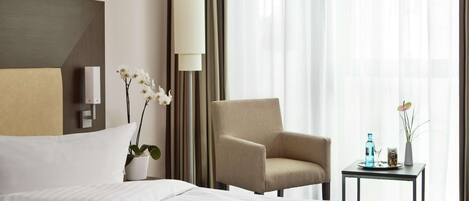 Habitación de hotel con cama, una silla, una mesita con un florero y una botella, y un ventanal con cortinas.