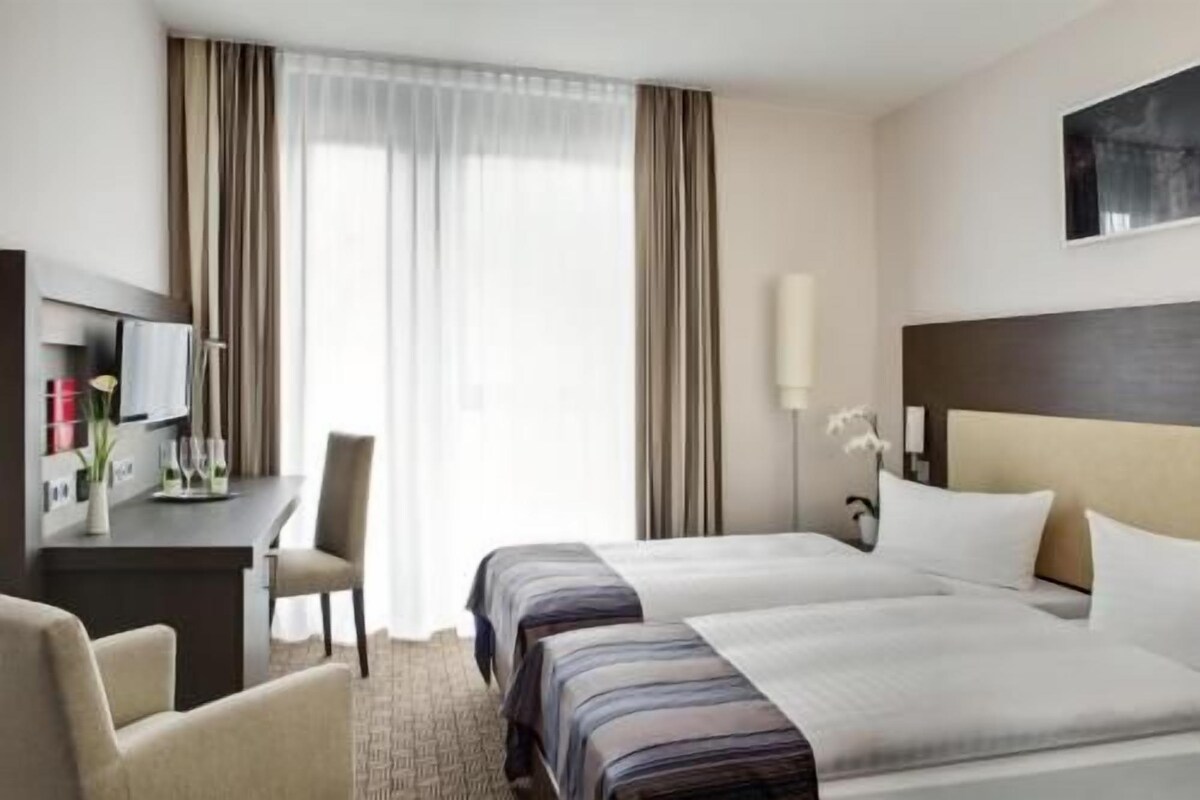Habitación de hotel con cama, escritorio, silla, televisor y ventana con cortinas.