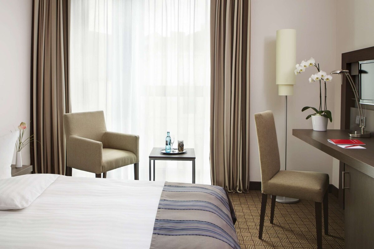 A hotel room with a bed, two armchairs, a small table with a bottle and glass, a desk with a lamp, and a potted plant.