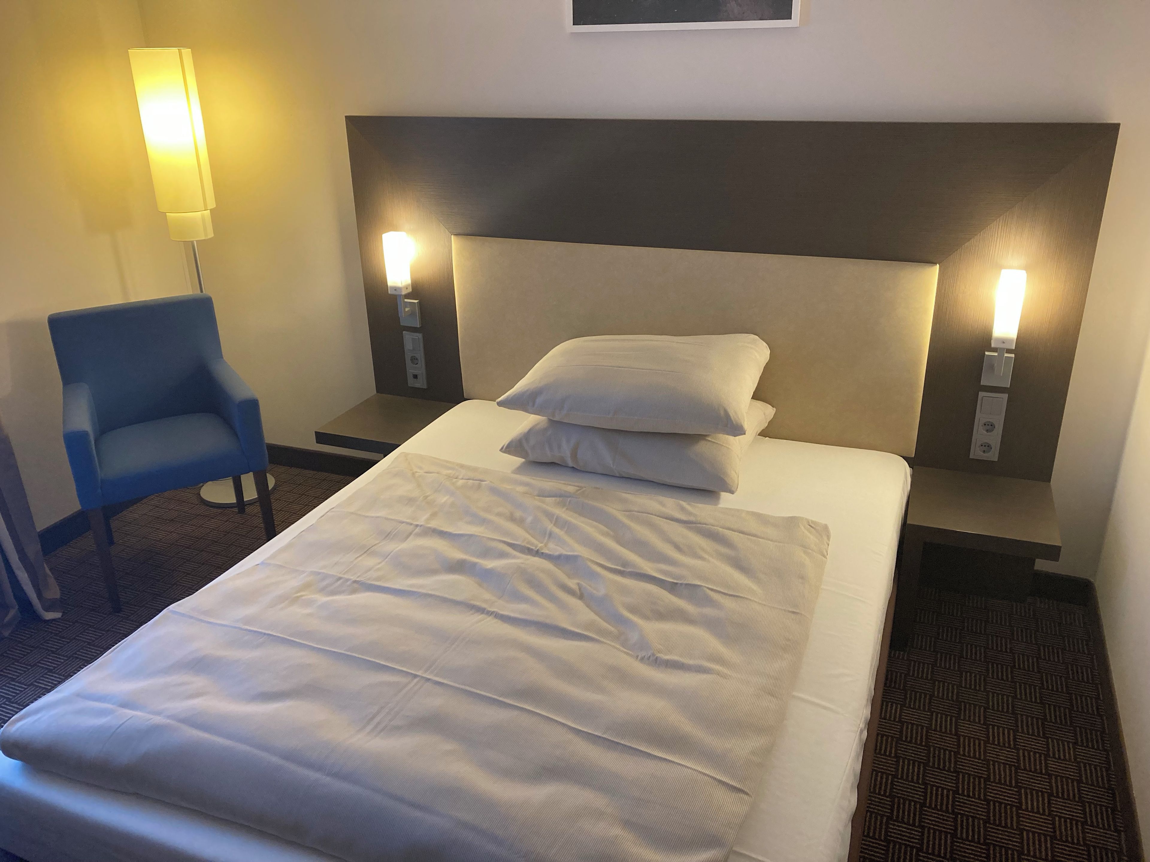 A hotel room with a bed, two bedside lamps, a chair, and a wall-mounted light fixture.