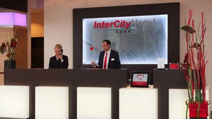 A reception desk at InterCity Hotel in Bonn with a digital screen displaying the hotel's name, a man speaking at the desk, and a potted plant to the right. - IntercityHotel Bonn (Bonn)