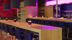 A modern bar with a long wooden counter, blue bar stools, and pendant lighting. - IntercityHotel Bonn (Bonn)