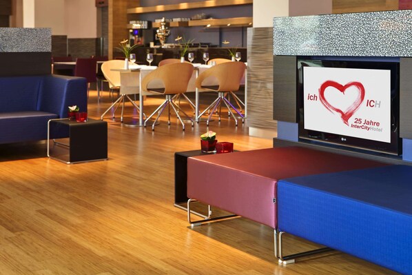 Un moderno vestíbulo de hotel con un área de estar, un televisor mostrando un logo de corazón, y un comedor al fondo.