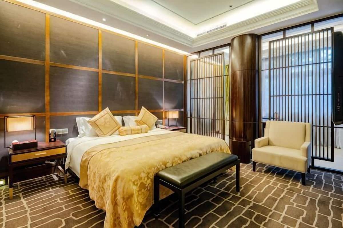 Habitación de hotel con una cama grande, un banco, un sofá, un escritorio con lámpara y una ventana grande con barrotes.
