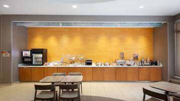 A modern kitchen area with a brick backsplash, equipped with a coffee machine, toaster, and various food items. There are wooden tables and chairs arranged for dining.