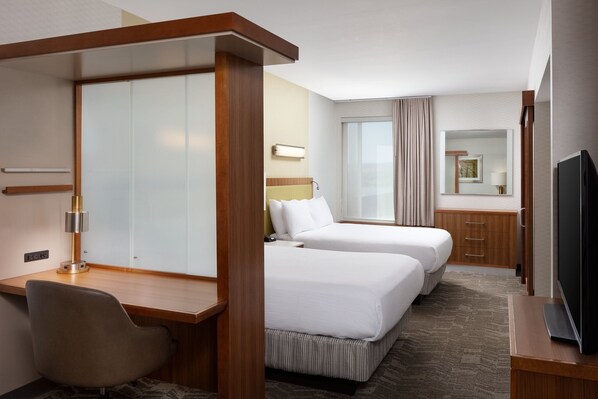 A hotel room with two beds, a desk, a chair, a TV, a mirror, and a window with curtains.