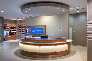 Zona de recepción de un hotel con un mostrador curvo, una pantalla digital que muestra opciones de habitaciones y un interior bien iluminado.