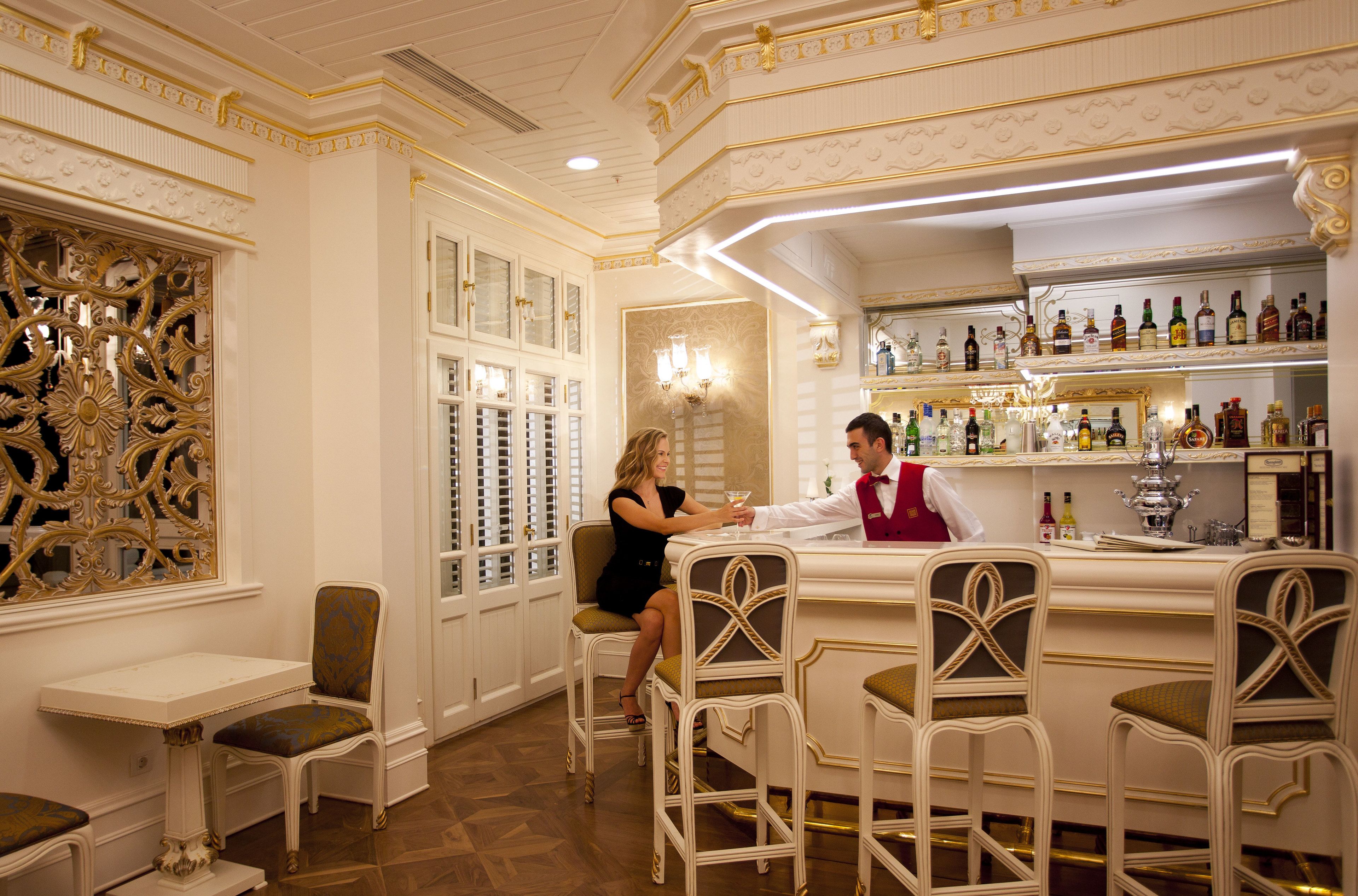 A bar area with ornate gold detailing, a bartender, and a woman seated at a table.