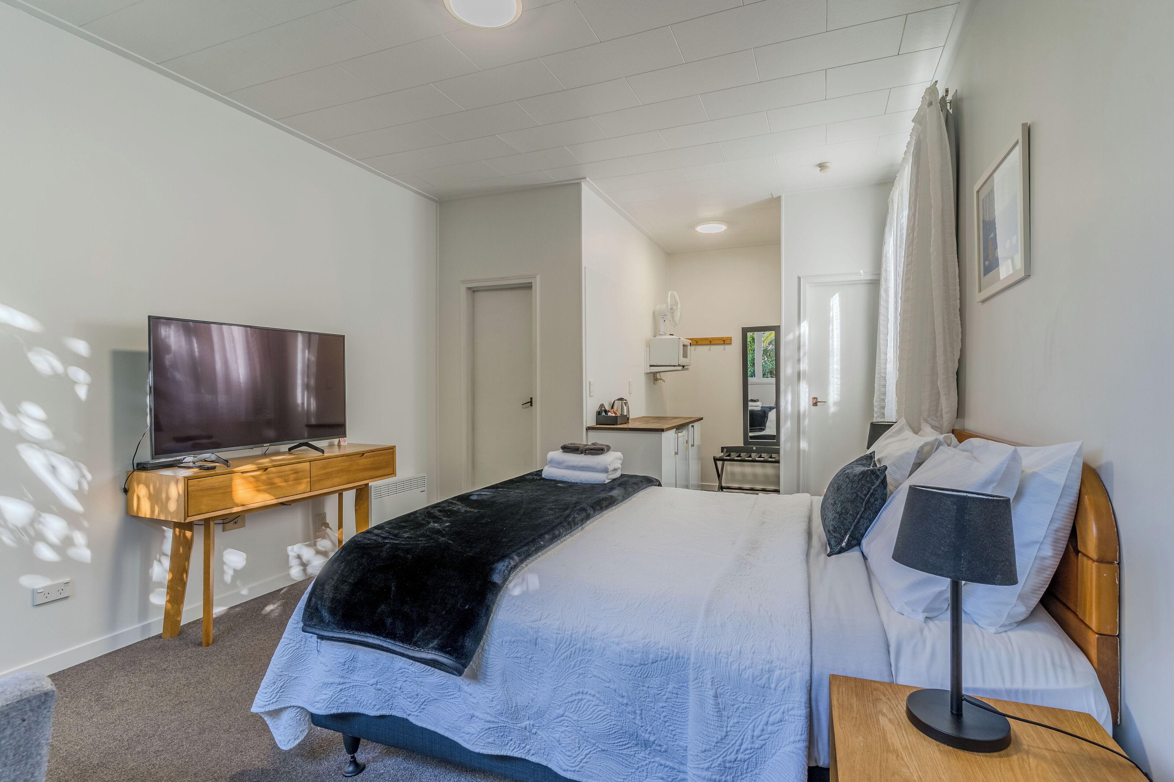 A hotel room with a bed, a desk with a television, a bedside table with a lamp, and a bathroom in the background.