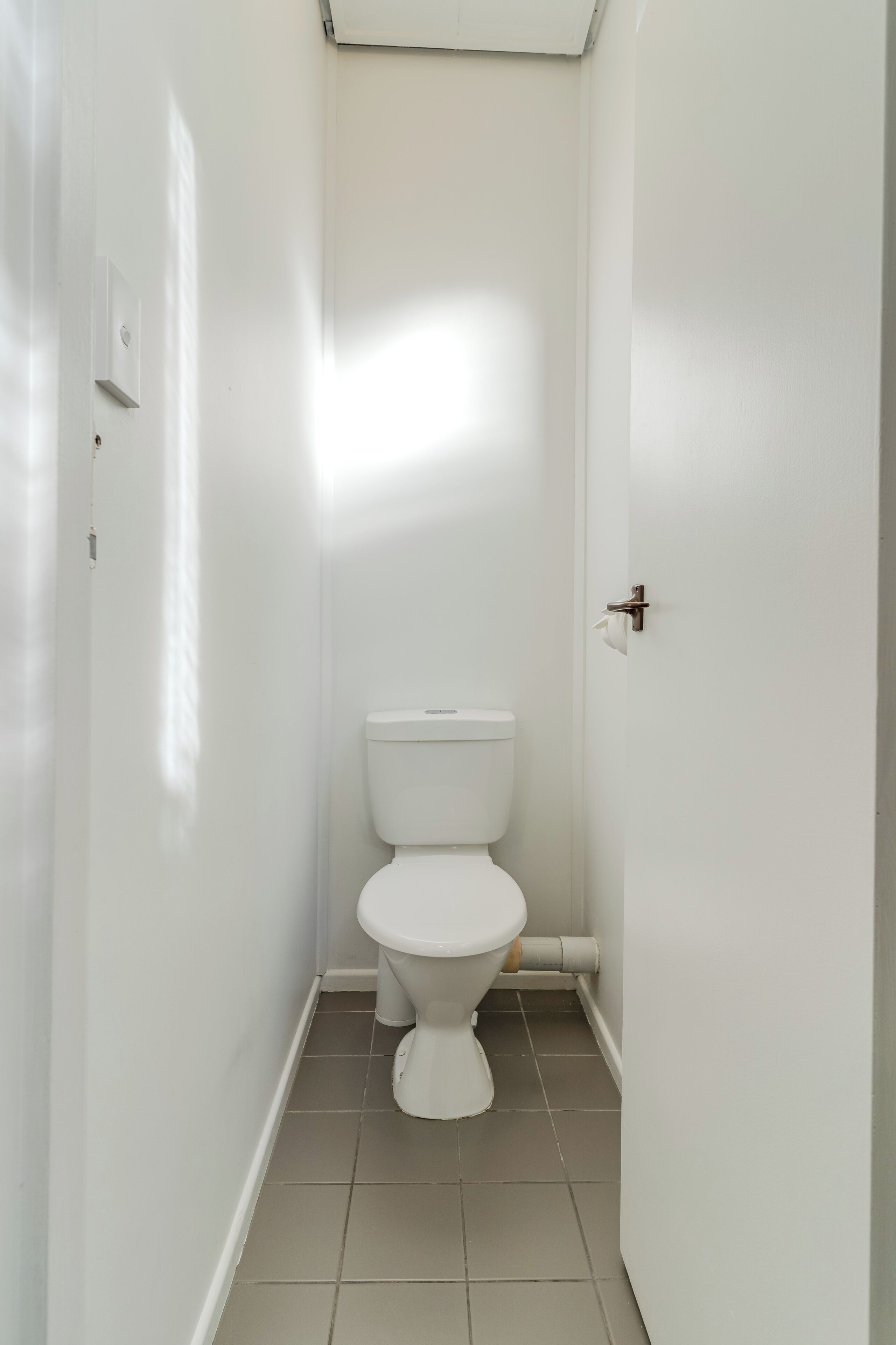 A small, white bathroom with a toilet, a white door, and a light switch on the wall.