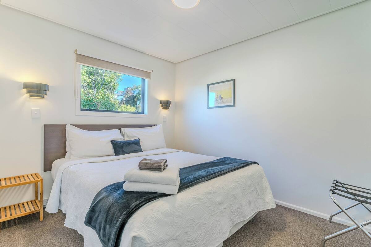 A neatly made bed with a white and dark blue blanket, a wooden nightstand, a wall-mounted light, and a window with a view of greenery.