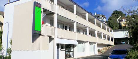 A multi-story hotel with a green sign, multiple balconies, and a parking area with a blue car.