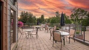 Patio esterno con tavoli e sedie, ringhiera in vetro e vista su alberi e un tramonto.